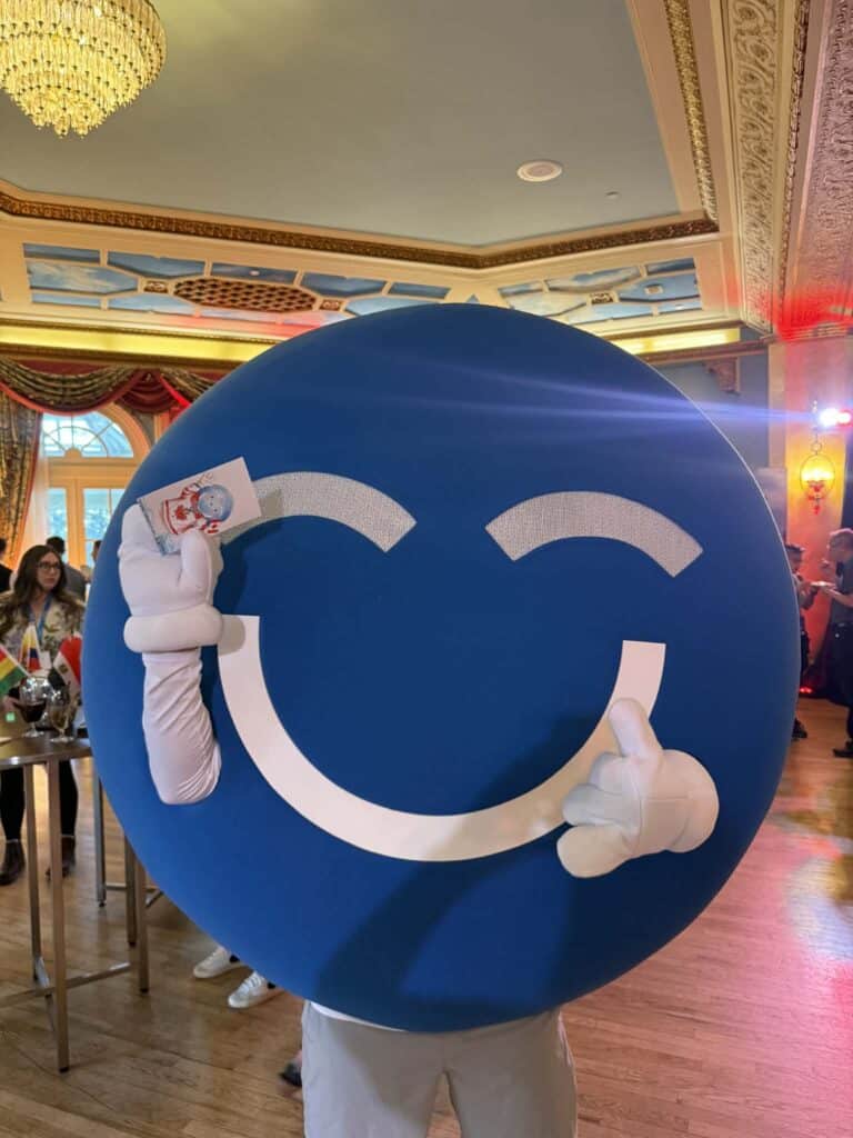 Blue circular mascot with a smiling face, holding a card, in a festive event setting, showcasing engaging brand activation at a corporate function.