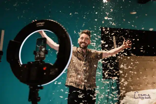 Man ppsing for a photo with a photo booth in front of a teal backdrop with golden confetti in the air