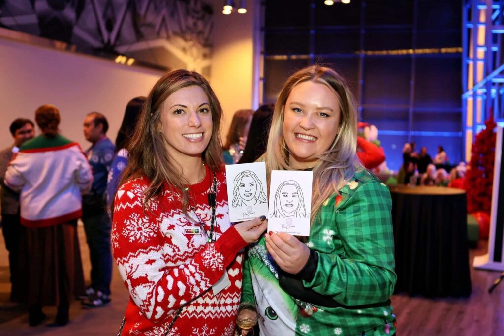 Two women smiling and holding illustrated portraits at a festive holiday event, surrounded by guests in holiday attire, showcasing joyful moments from a photo booth experience.